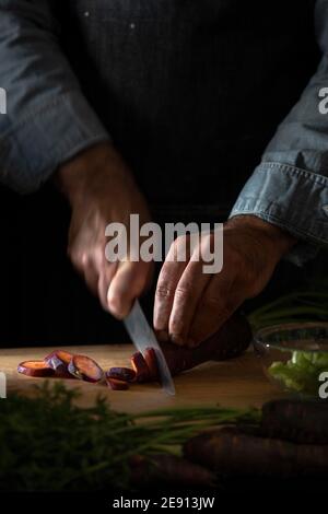 A closeup of a chef chopping a purple carrot on a wooden cutting board. Stock Photo