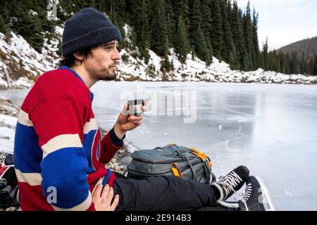 Young hockey player resting near frozen lake and drinking hot beverage Stock Photo