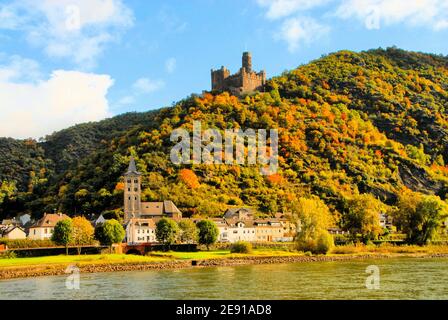 A castle stands against a blue sky with white clouds on an autumn hilltop in the Rhine Gorge, while a small town is below beside the Rhine River. Stock Photo