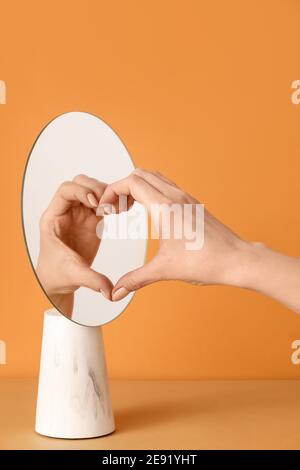 Female hand and its reflection in mirror making heart against color background Stock Photo
