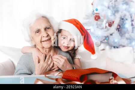 Smiling little granddaughter in santa hat sit hugging with grandma at Christmas Stock Photo