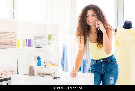 Cute tailor making notes talking on phone in sewing studio Stock Photo
