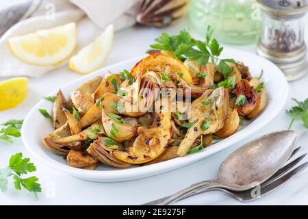 Sauteed artichokes with fresh parsley, roarted lemon slices and garlic served on a white oval plate Stock Photo