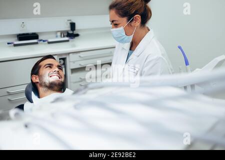 Female dentist in protective mask while treating a patient in dental office. Dental doctor talking with a smiling male patient sitting in dentist's ch Stock Photo