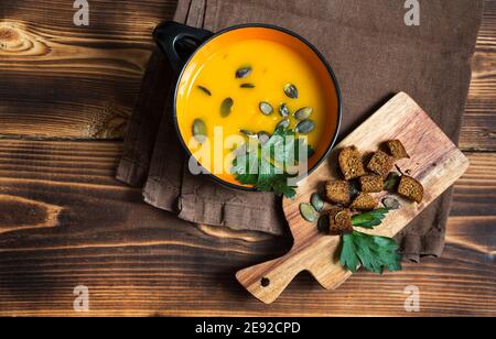 Pumpkin soup in a bowl with seeds and parsley on a wooden background. Crackers with spices on the board. Copy space Stock Photo