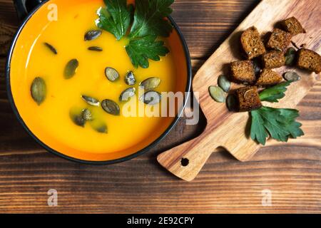 Pumpkin soup in a bowl with seeds and parsley on a wooden background. Crackers with spices on the board. Copy space Stock Photo
