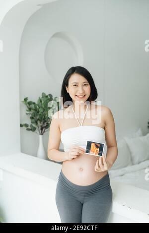 Pregnant woman holds ultrasound photo on the belly in living room Stock Photo