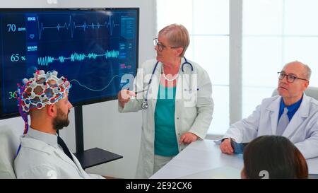 Scientist doing brain analysis using headset with sensors on doctor in hospital meeting room. Monitor shows modern brain study while team of scientist adjusts the device working in conference office Stock Photo
