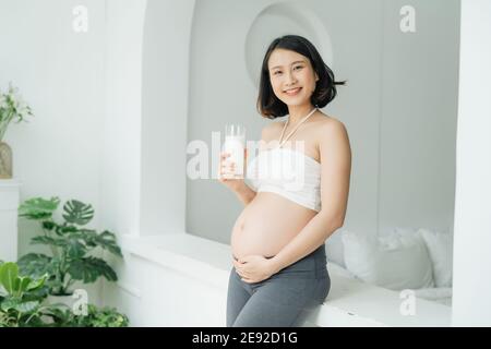 Happy pregnant woman drinking a glass of fresh milk and touching her belly with care by the window in bedroom Stock Photo