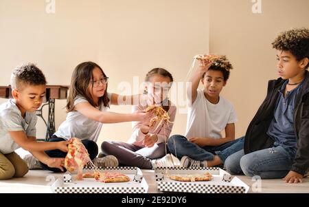 Five multiethnic pretty children little boys and girls sit indoor eating pizza italian fast food cuisine. Alpha generation share slices, spend time to Stock Photo