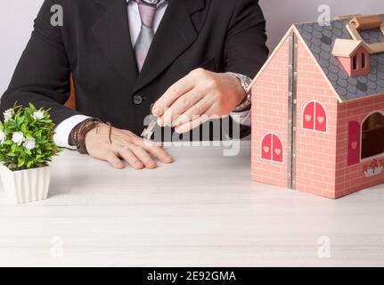 Image of an office table with a wooden house and an artificial plant with a real estate consultant handing over the key to a property. Stock Photo