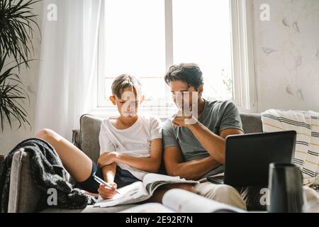 Son looking in book while father sitting by with laptop on sofa at home Stock Photo