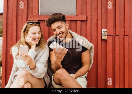 Friends laughing while using mobile phone against cottage Stock Photo