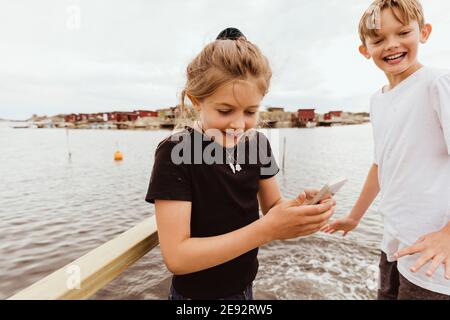 Sister with smart phone standing by smiling brother against sea Stock Photo
