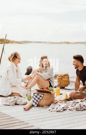 Happy young friends spending leisure time while sitting on jetty against sky and sea Stock Photo