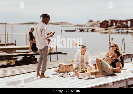 Young man using phone standing by friends on jetty during sunny day Stock Photo