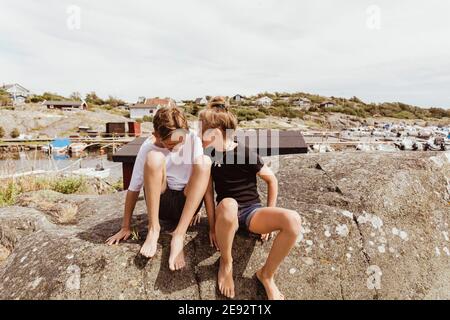 Happy siblings sitting on rock against sky during summer vacation Stock Photo