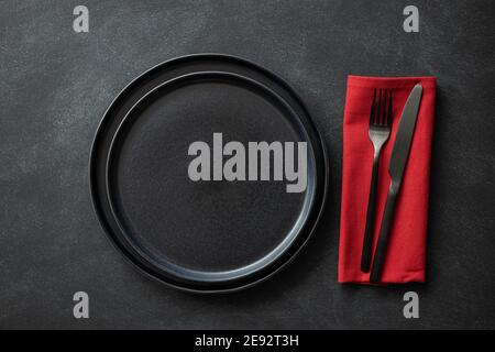 Empty black plates with black cutlery and red napkin on black table. Top view. Stock Photo