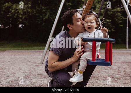 Smiling father kissing son while sitting on swing in playground Stock Photo