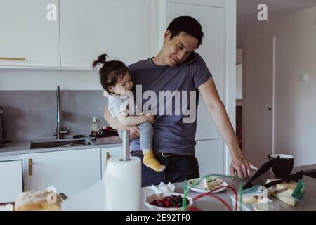 Father talking on smart phone while using digital tablet while carrying baby son in kitchen Stock Photo
