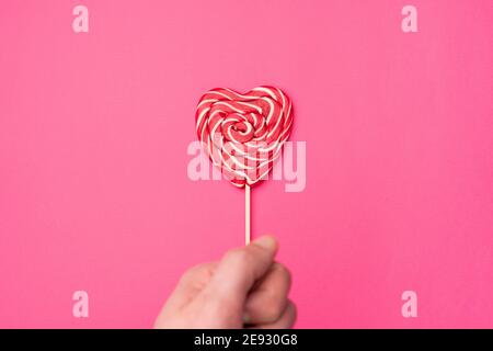 Handholds red heart-shaped lollipop candy isolated on an empty colorful pink background. Symbol of love for Happy Women's, Valentine's Day, Birthday greeting romantic postcard. Flat lay, top view, copy space Stock Photo