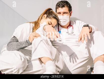 Tired doctors in medical masks and uniform leaning on each other while sleeping on balcony after hard work during COVID 19 pandemic Stock Photo