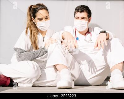 Tired doctors in medical masks and uniform leaning on each other while sleeping on balcony after hard work during COVID 19 pandemic Stock Photo