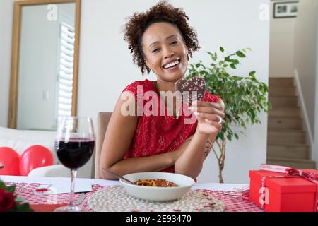 Portrait of african american woman holding heart shaped cookie on videocall at home Stock Photo