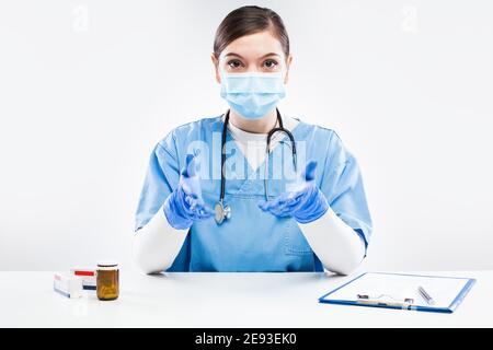 Young female UK NHS doctor wearing protective glove and face mask,talking online to patient,young GP woman in blue uniform consulting person remotely, Stock Photo