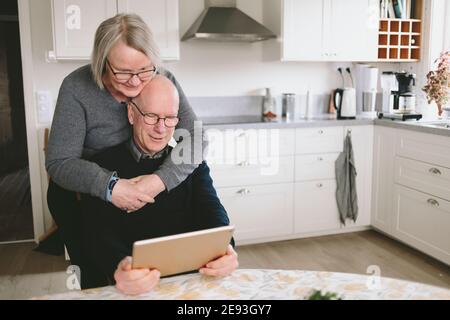 Smiling couple embracing and using tablet at kitchen table Stock Photo