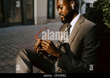Male entrepreneur looking at eyeglasses while sitting on footpath Stock Photo