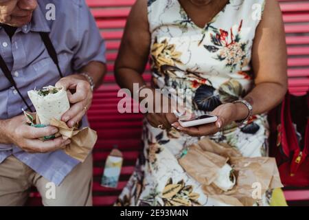 Senior man with food sitting by woman using smart phone on bench Stock Photo