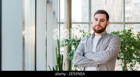 Caucasian man standing near full length window in modern office with plants. Successful bearded young man crossed arms. Handsome businessman smart man Stock Photo