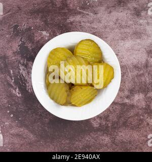 Top view of a small white bowl filled with hamburger dill pickle slices on a maroon tabletop illuminated with natural lighting. Stock Photo