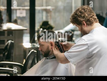 Close up master barber, stylist does the hairstyle to guy, young man. Professional occupation, male beauty concept. Cares of hair, mustache, beard of client. Soft colors and focus, vintage. Stock Photo