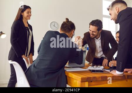 Two happy young businessmen shaking hands in office with smiling coworkers watching Stock Photo