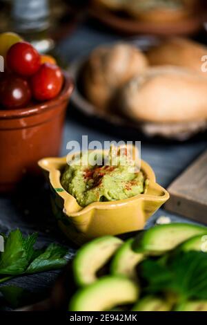closeup of a ceramic bowl with guacamole, next some slices of avocado in a plate and some cherry tomatoes in an earthenware bowl on a gray rustic wood Stock Photo