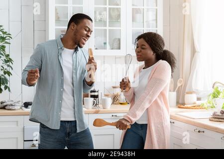Funny Black Couple Singing And Dancing In Kitchen, Using Utensils As Microphones Stock Photo