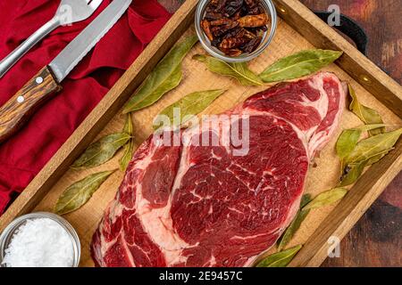 Raw beef ribeye on a round slate plate with glass containers containing peppercorns, coarse salt and cayenne chillies on an old wooden table in brown Stock Photo