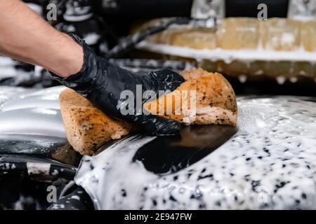 Close up of car wash worker wearing protective gloves and washing car engine with soapy sponge. Cleaning services. Stock Photo