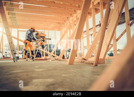 Caucasian Contractor Carpenter Worker in His 40s Using Commercial Grade Circular Saw in Construction Zone. Industrial Theme. Wooden Skeleton Framing B Stock Photo