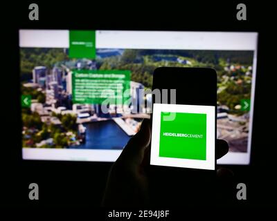 Person holding smartphone with logo of German building materials company HeidelbergCement AG on display in front of website. Focus on phone screen. Stock Photo