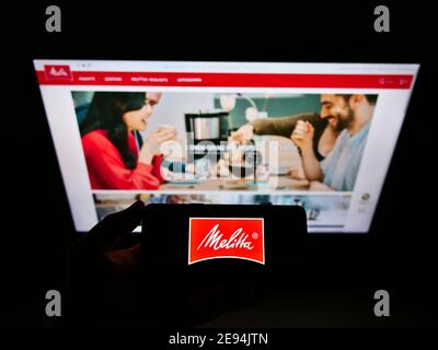 Person holding smartphone with logo of German coffee retail company Melitta Unternehmensgruppe Bentz KG on display. Focus on mobile phone screen. Stock Photo