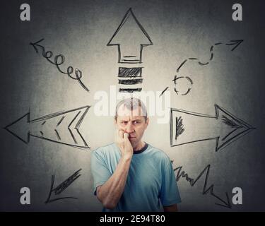 Skeptical thoughtful looking middle aged man, keeps hand under chin, puzzled suspicious expression, has to choose a direction from different arrows po Stock Photo