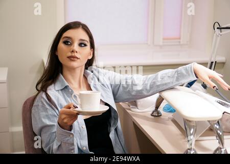 Beautiful woman does a manicure in a beauty salon. She holds a white cup of coffee in her hand. Stock Photo
