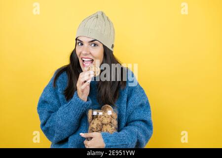 Young beautiful woman wearing blue casual sweater and wool hat holding chocolate chips cookies jar very happy, taking a cookie from the jar and eating Stock Photo