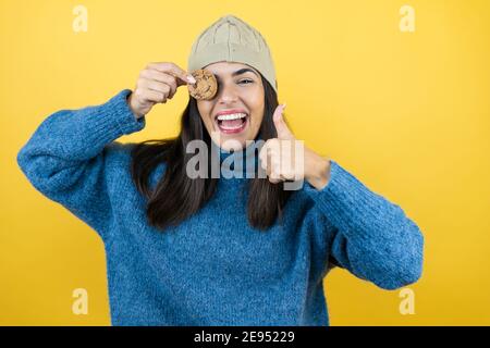 Young beautiful woman wearing blue casual sweater and wool hat holding healthy chocolate cookie over eye happy with big smile doing ok sign, thumb up Stock Photo