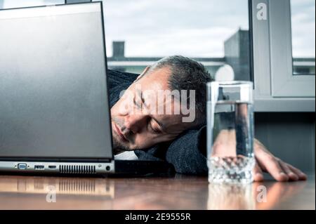 Tired business man sleeps near his laptop. Young bearded tired man sleeping on office table. Work routine concept. Stock Photo