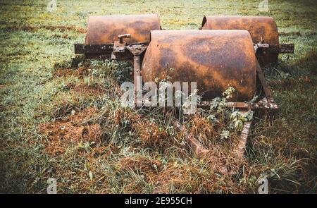 Vintage rusty agricultural field roller abandoned on overgrown green grass in rural countryside. Stock Photo