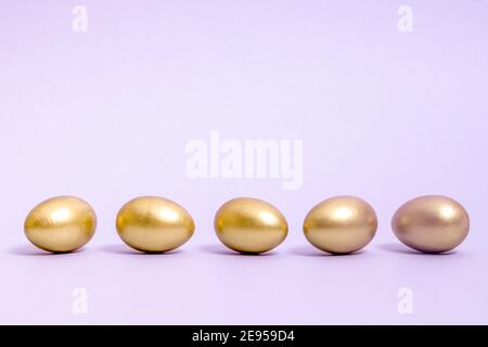 Five Easter eggs decorated with gold paint lie in a row against a lavender background. Concept for Easter, spring. Selective focus. Copy space. Stock Photo